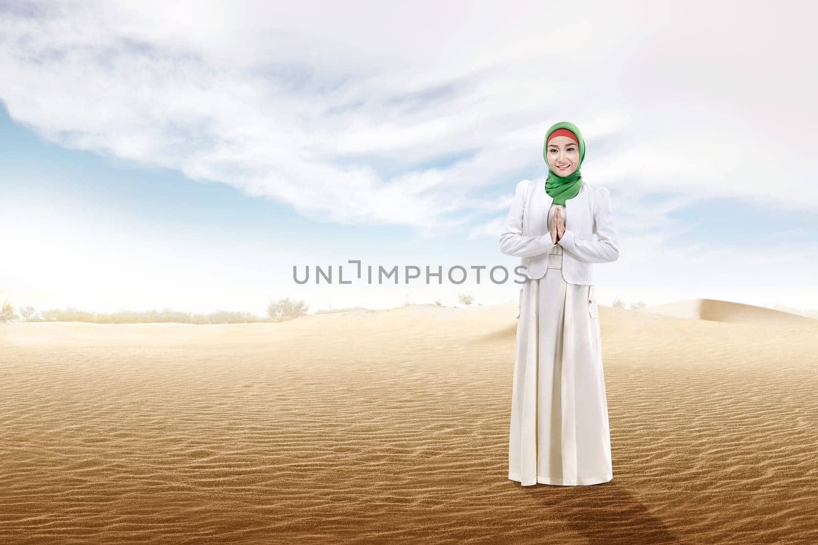 Smiling asian muslim woman in veil praying on the sand dune with blue sky background