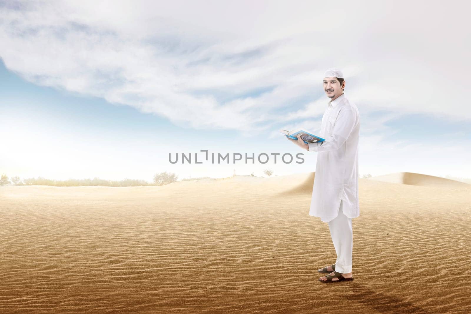 Happy asian muslim man holding the open quran standing on the sand dune with blue sky background