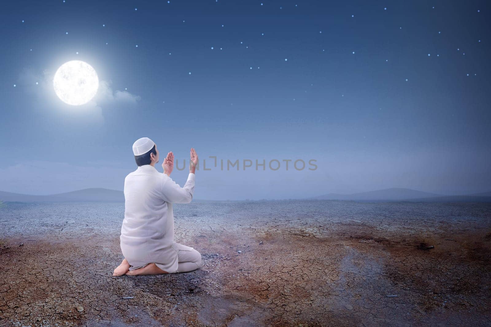 Rear view of asian muslim man sitting in pray position while raised hands and praying on the sand dune with moon and night scene background