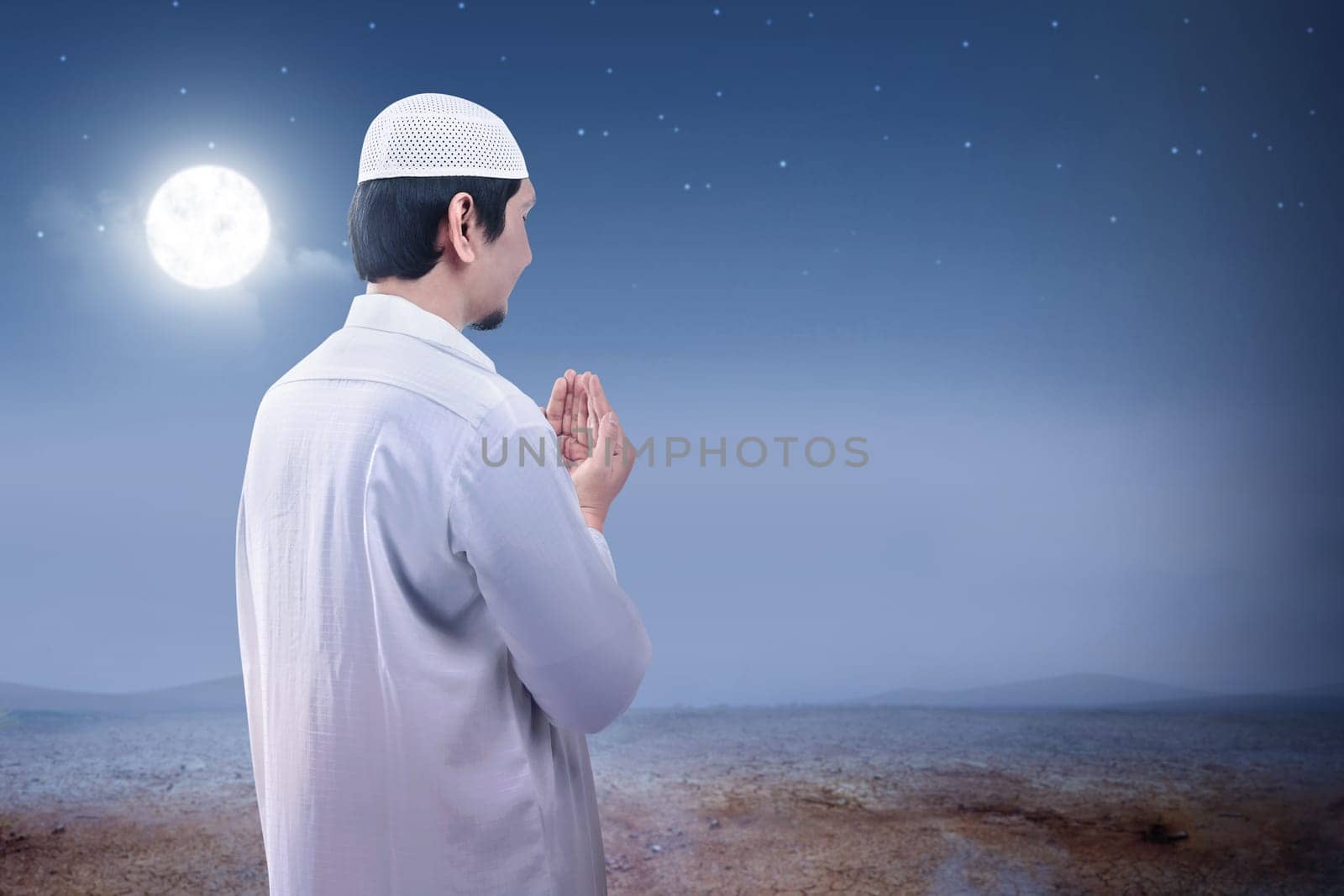 Rear view of asian muslim man raised hands and praying on the sand dune with moon and night scene background