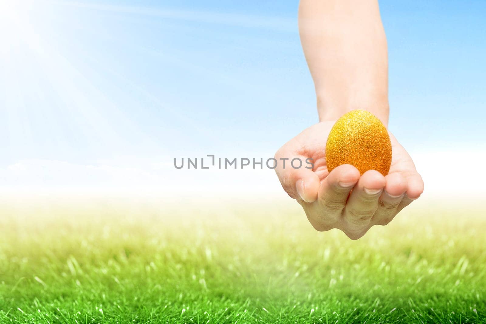 Human hand holding yellow easter egg on the grass field with sunlight and blue sky background. Happy Easter