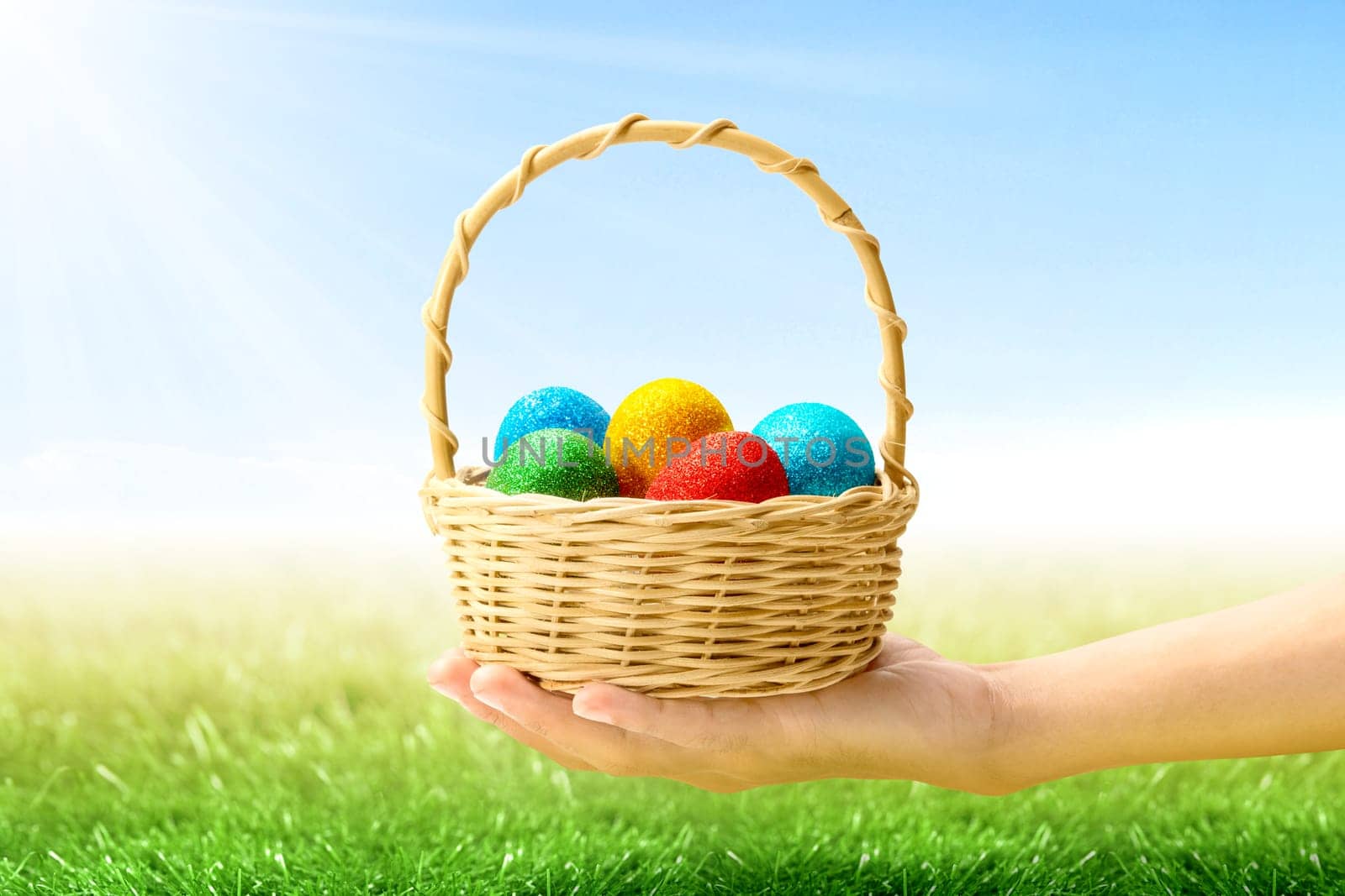 Human hand holding wooden basket with colorful easter eggs on the grass field with sunlight and blue sky background. Happy Easter