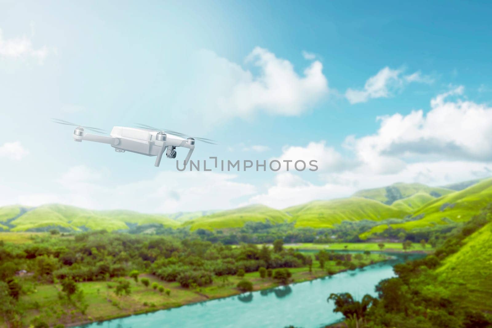 White drone with camera flying over green hills with river and trees over blue sky background