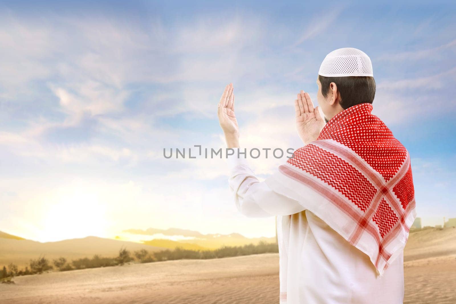 Rear view of asian muslim man with cap and turban standing and praying with raised arms on the sand with blue sky background