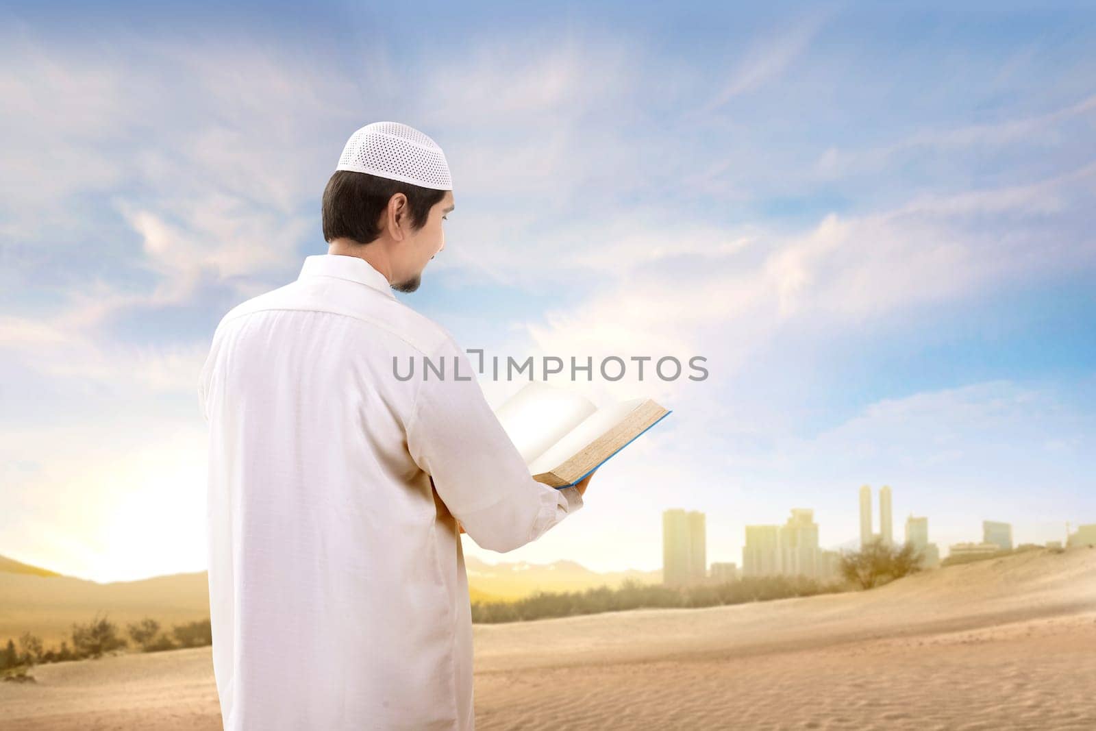 Rear view of asian muslim man with cap standing and reading the quran on the sand with blue sky background
