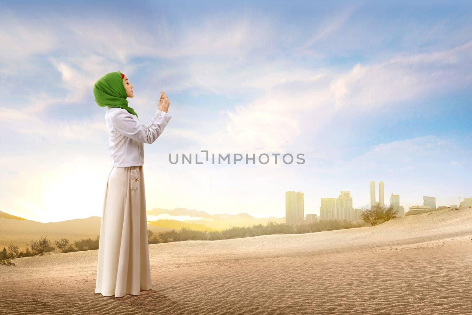 Pretty asian muslim woman in veil standing and praying with raised arms on the sand with blue sky background