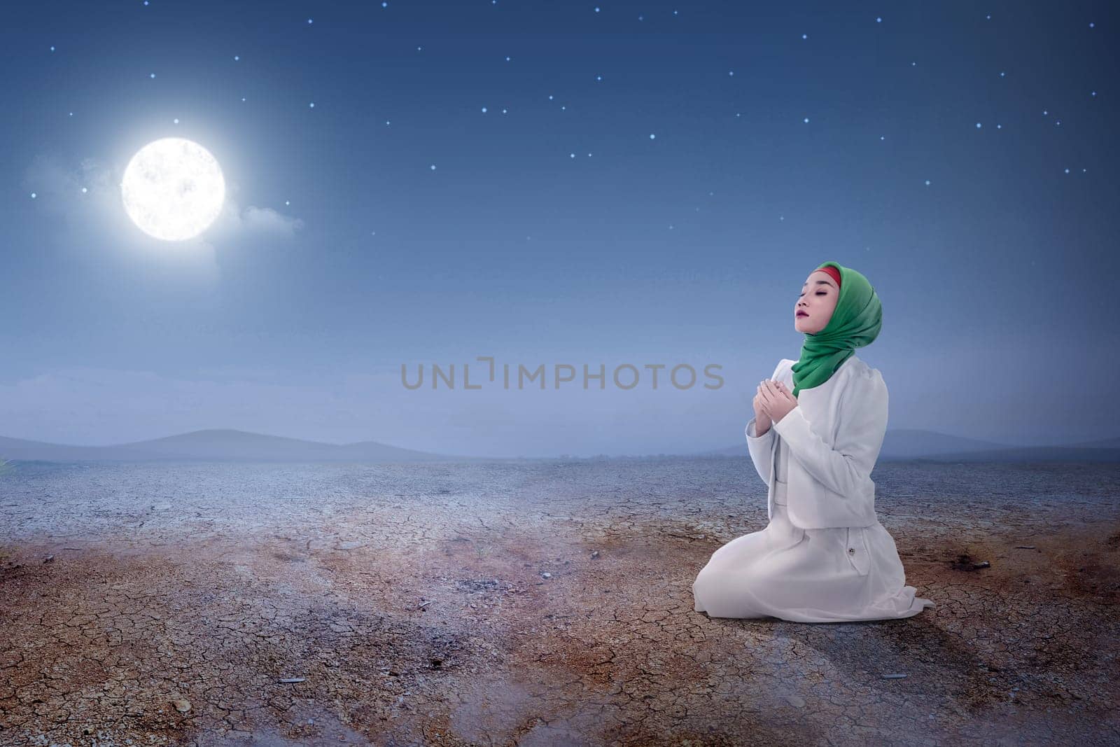 Young asian muslim woman sitting in pray position while raised hands and praying on the sand dune with moon and night scene background