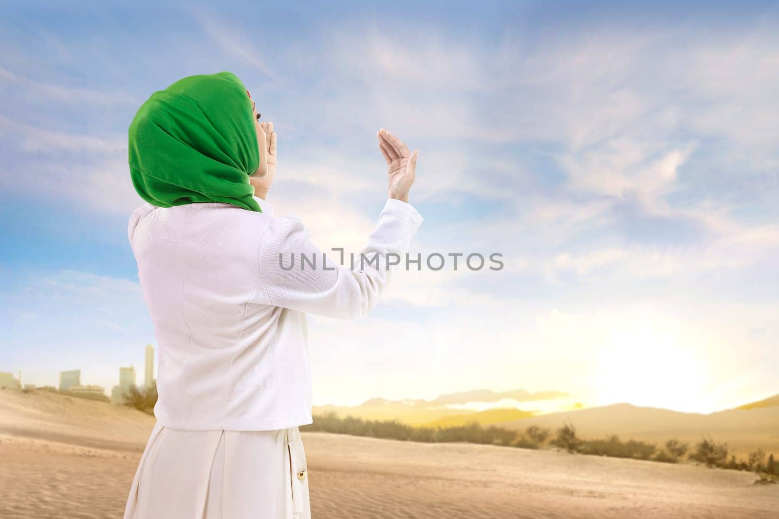 Beautiful asian muslim woman in veil standing and praying with raised arms on the sand with blue sky background