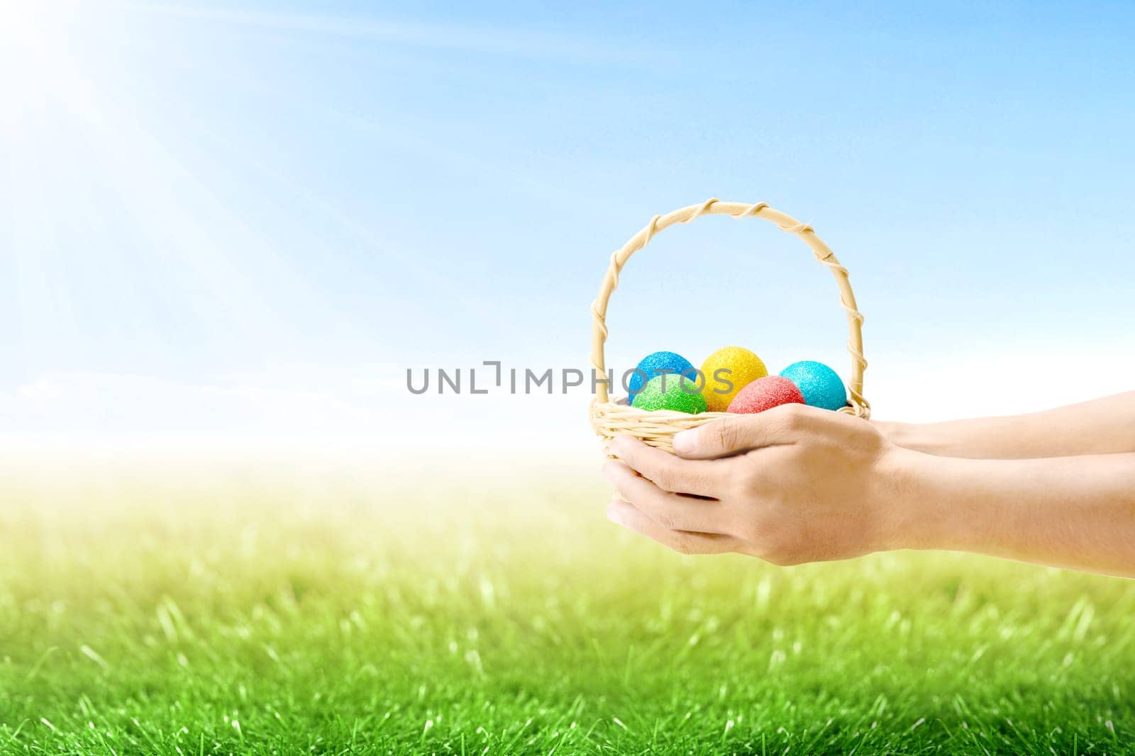 Human hands holding wooden basket with colorful easter eggs on the grass field with sunlight and blue sky background. Happy Easter