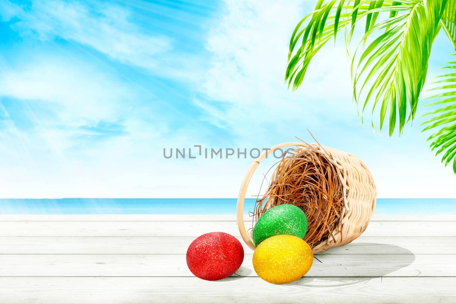 Colorful easter eggs spilled from nest on wooden basket to wooden table with palm branch and ocean view over blue sky background. Happy Easter