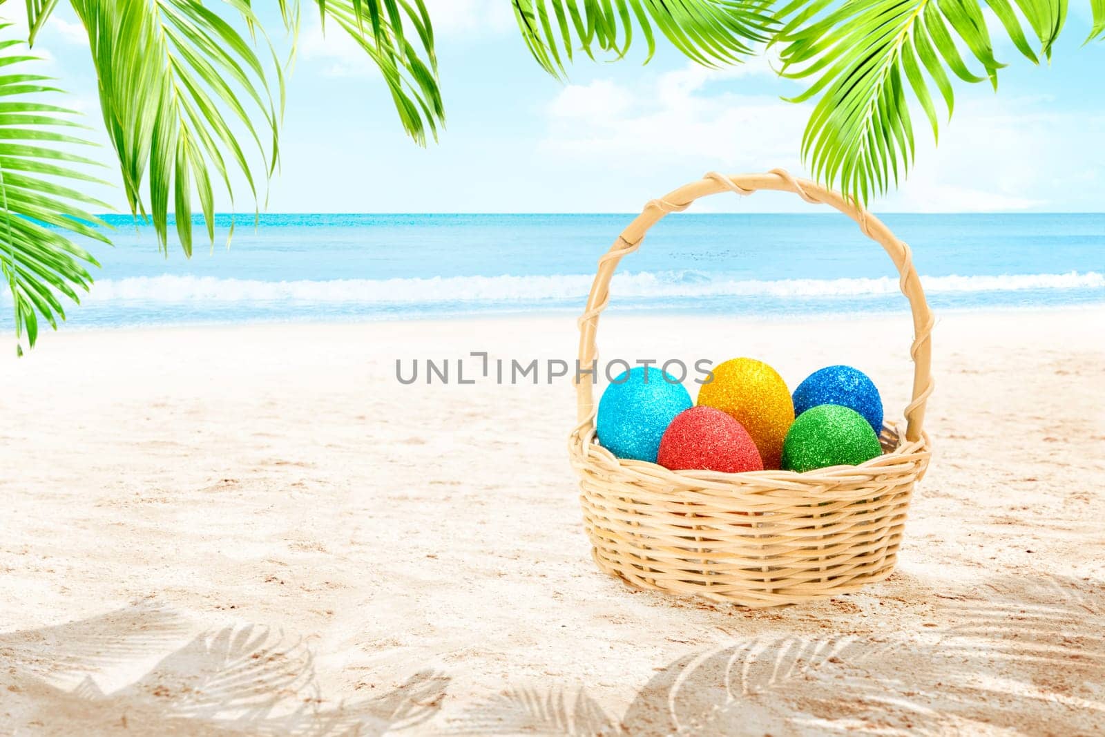 Colorful easter eggs in wooden basket on the sandy beach with palm branch and waving sea over blue sky background. Happy Easter