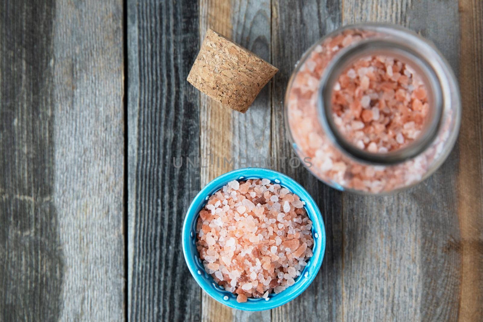 Pink Himalayan salt crystals in glass bottle and small bowl on wooden table flat lay.