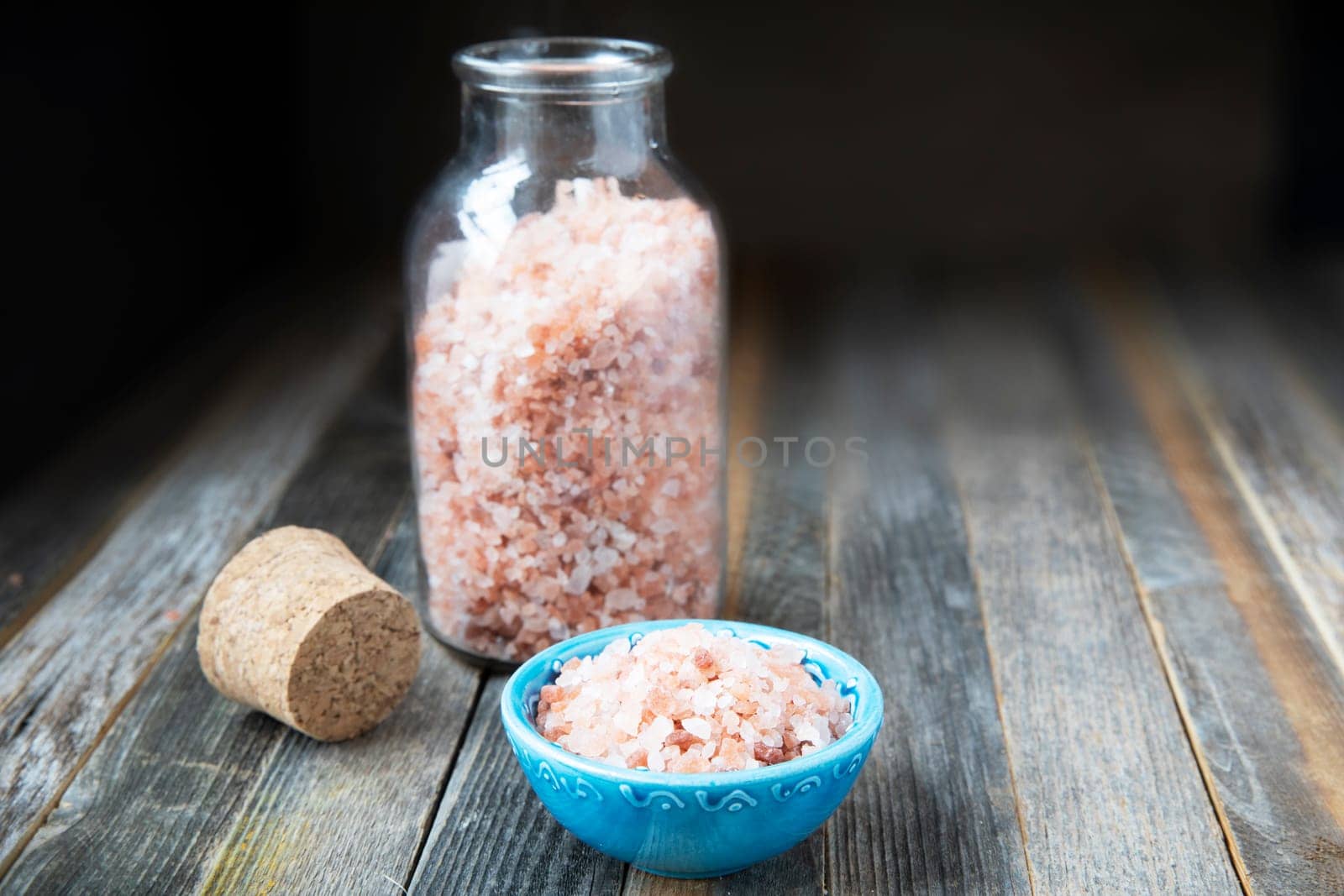 Pink Himalayan salt crystals in glass bottle and small bowl on wooden table.