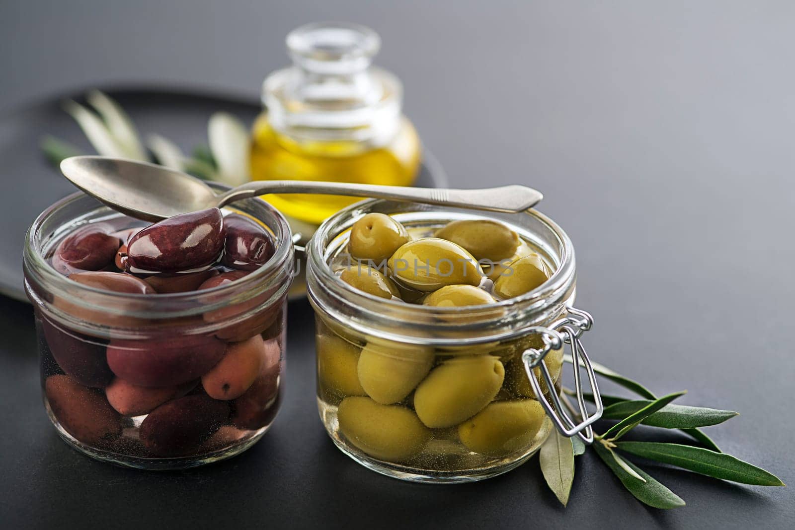 Pickled green and black olives in glass jar with branch leaves close up