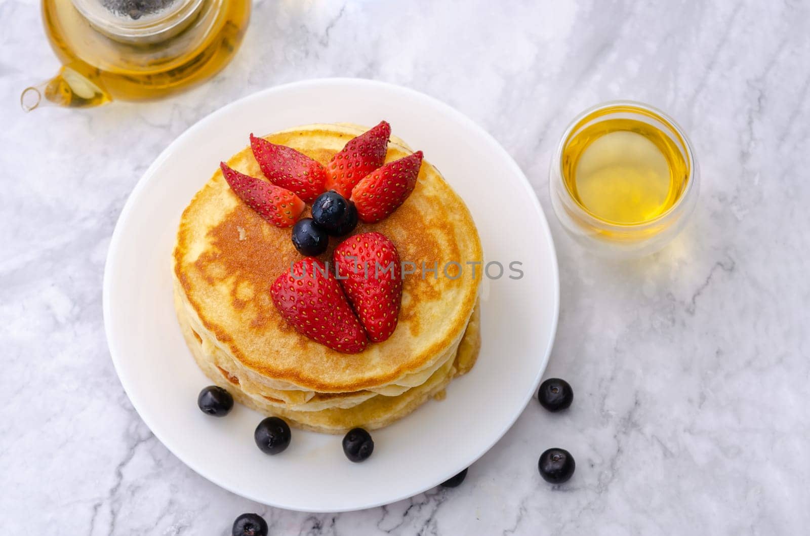 Tasty pancakes with fresh strawberry and blueberry on white plate with teapot on marble background. Table top view