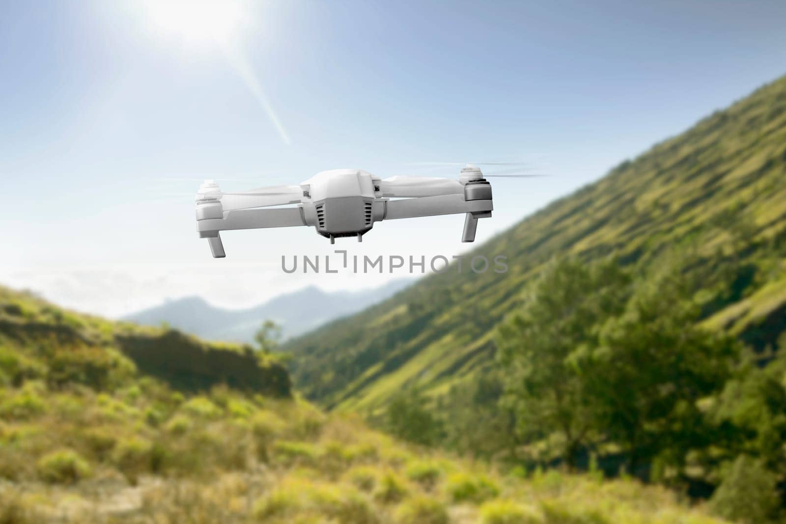 White drone with camera flying above the mountain with green grass and trees with sunlight and blue sky background