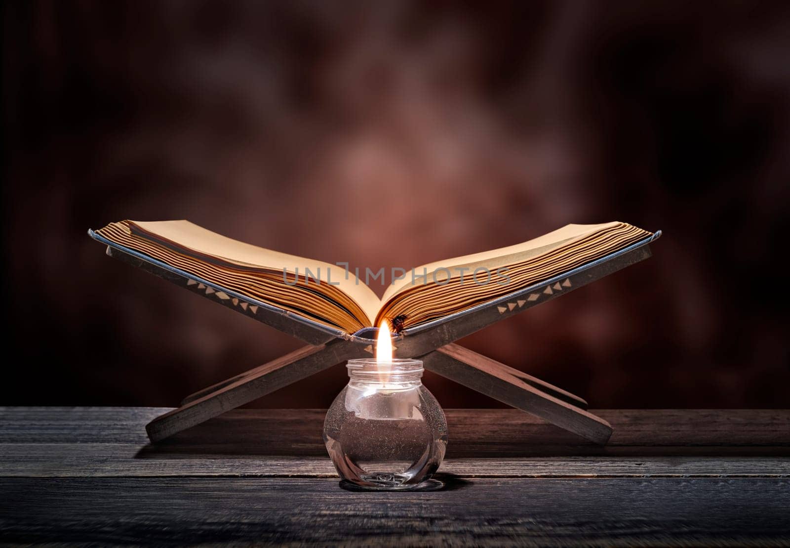 Quran open in wooden placemat and candle on wooden table over blurry background