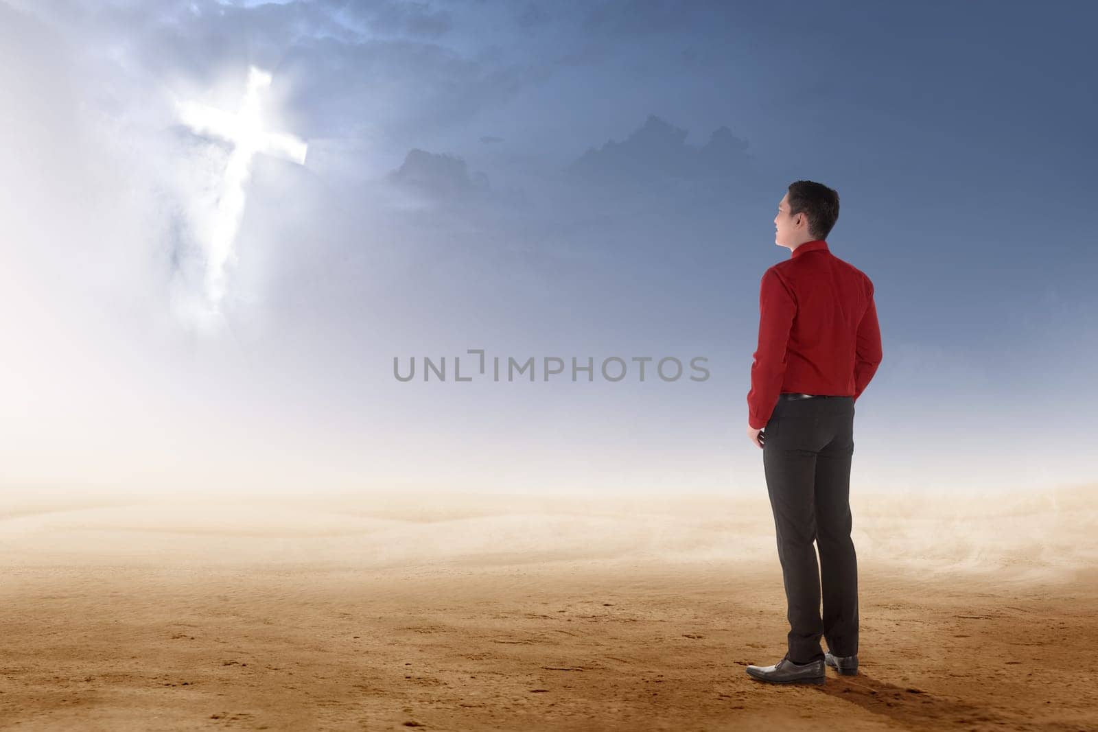 Rear view of asian businessman standing on desert and looking at glowing christian cross on the sky
