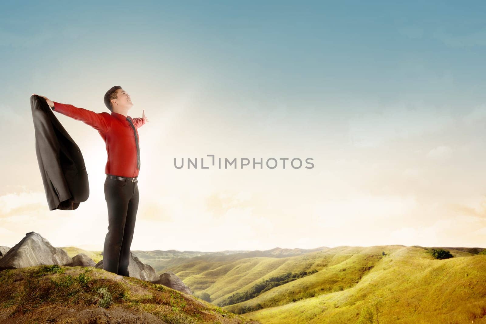 Happy asian businessman holding a suit standing on top of mountain celebrate his successful over blue sky background