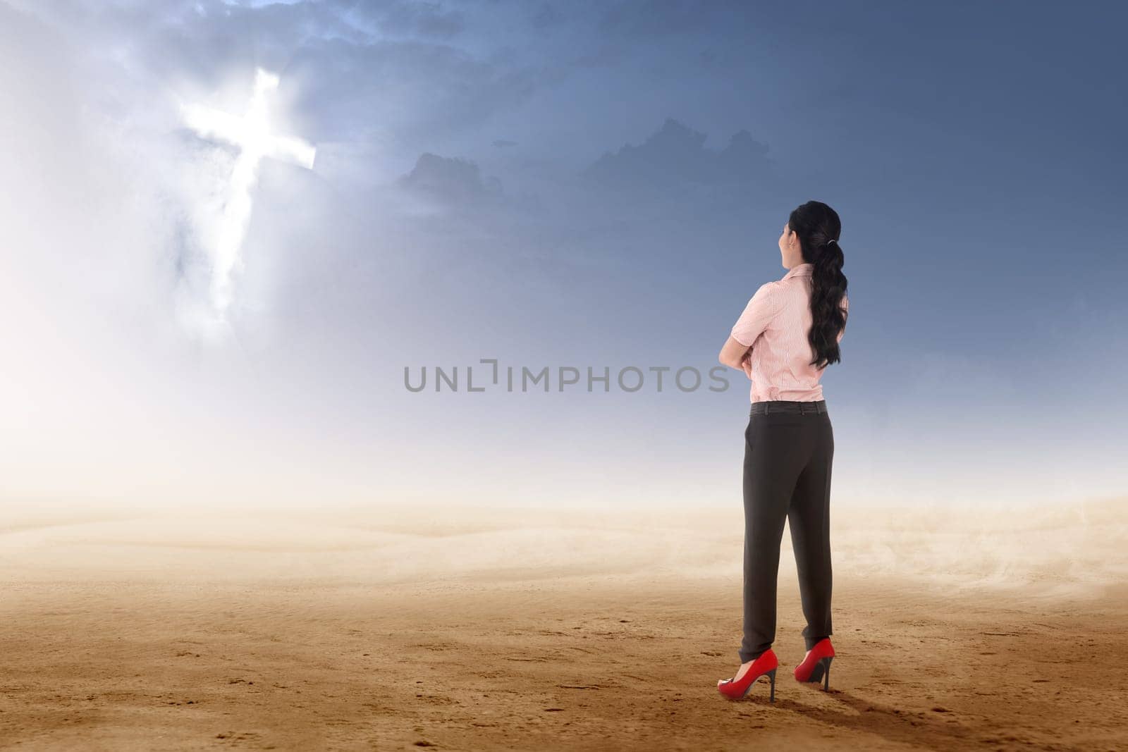 Rear view of asian businesswoman standing on desert and looking at glowing christian cross on the sky