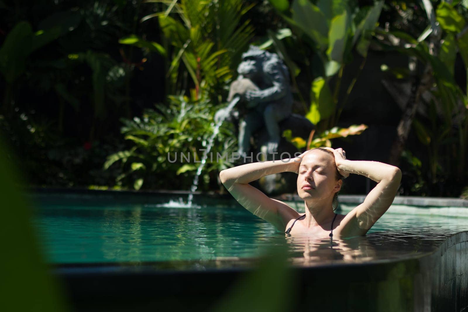 Sensual young woman relaxing in outdoor spa infinity swimming pool surrounded with lush tropical greenery of Ubud, Bali. Wellness, natural beauty and body care concept.