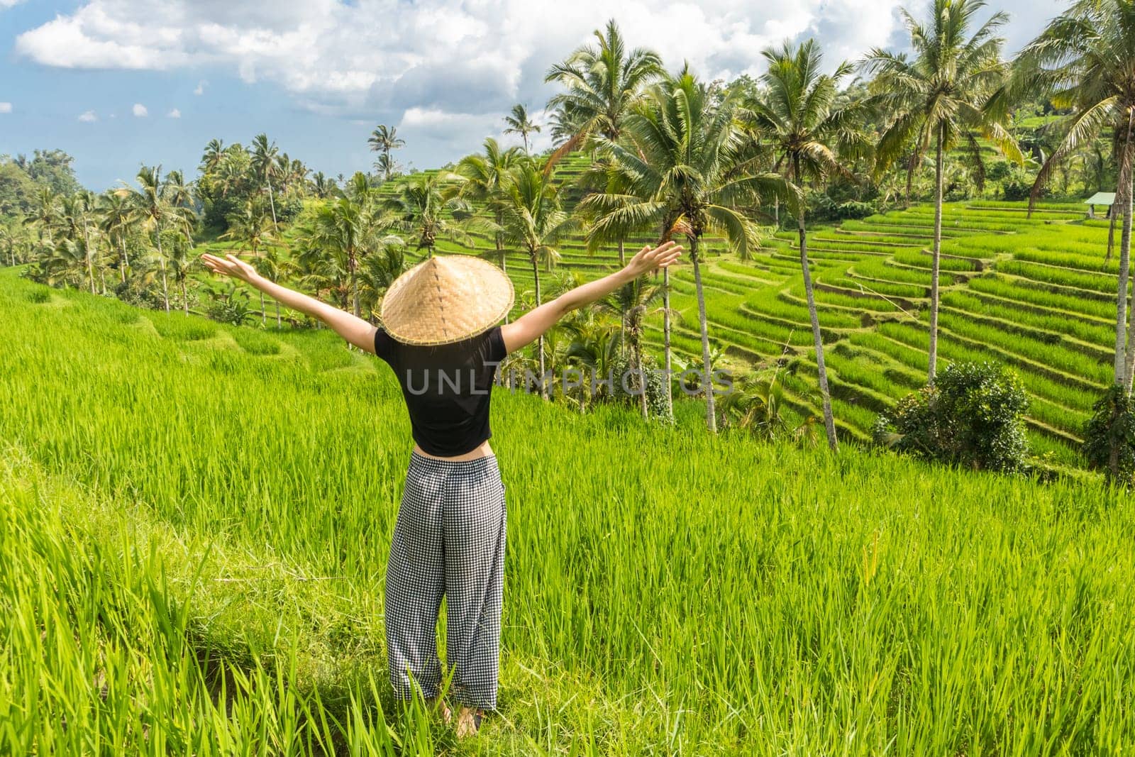 Relaxed fashionable female traveler wearing small backpack and traditional asian paddy hat, arms rised to sky, enjoying pure nature at beautiful green rice fields and terraces on Bali island.