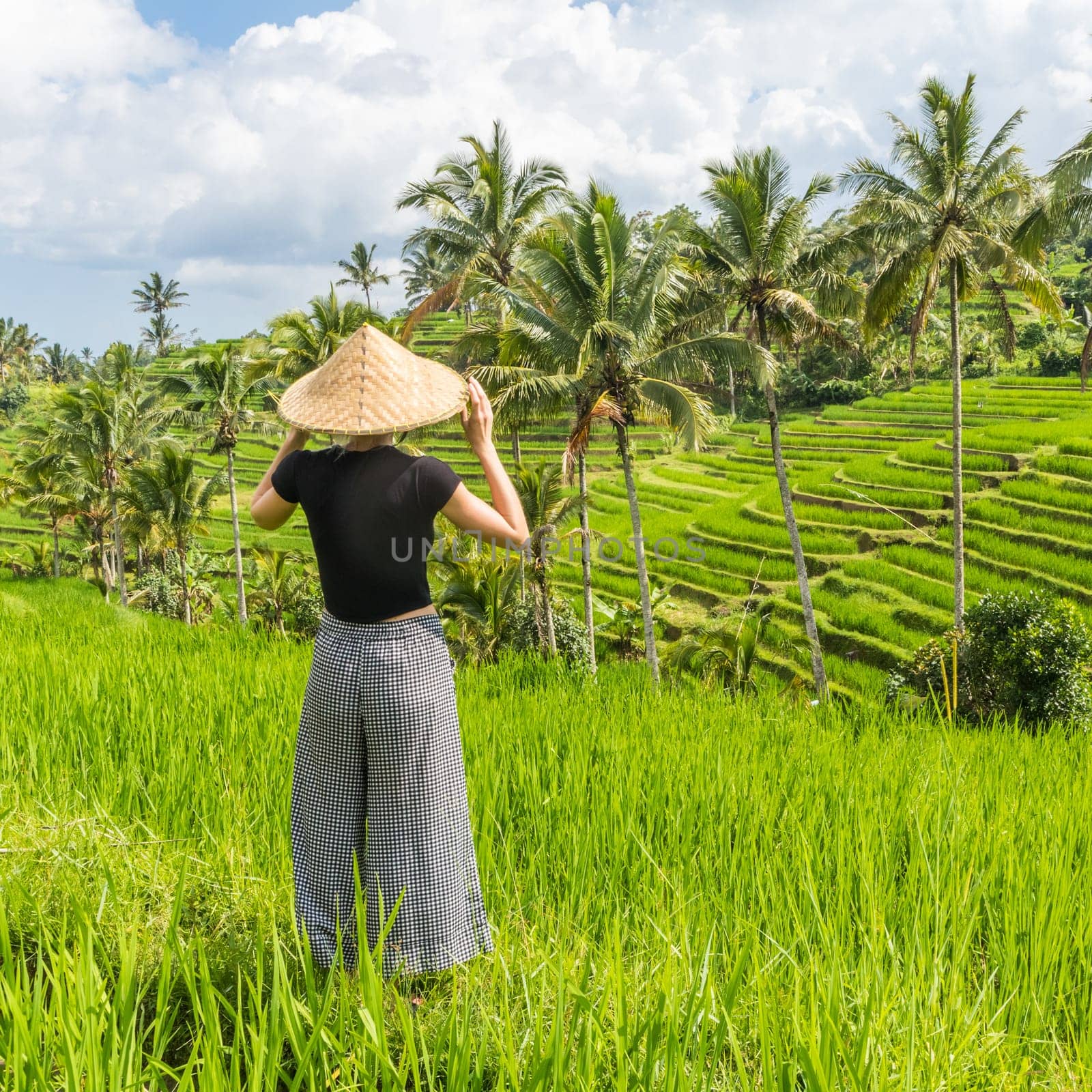 Relaxed fashionable caucasian female tourist wearing small backpack and traditional asian paddy hat looking at beautiful green rice fields and terraces on Bali island.