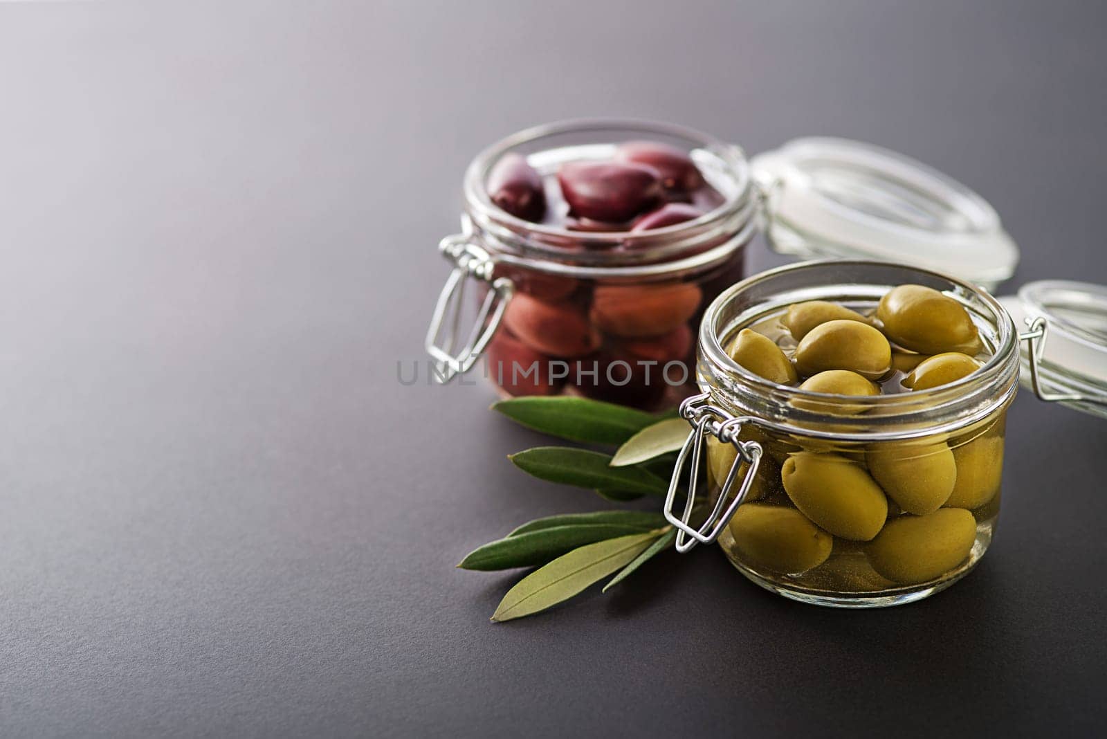 Pickled green and black olives in glass jar with branch leaves close up