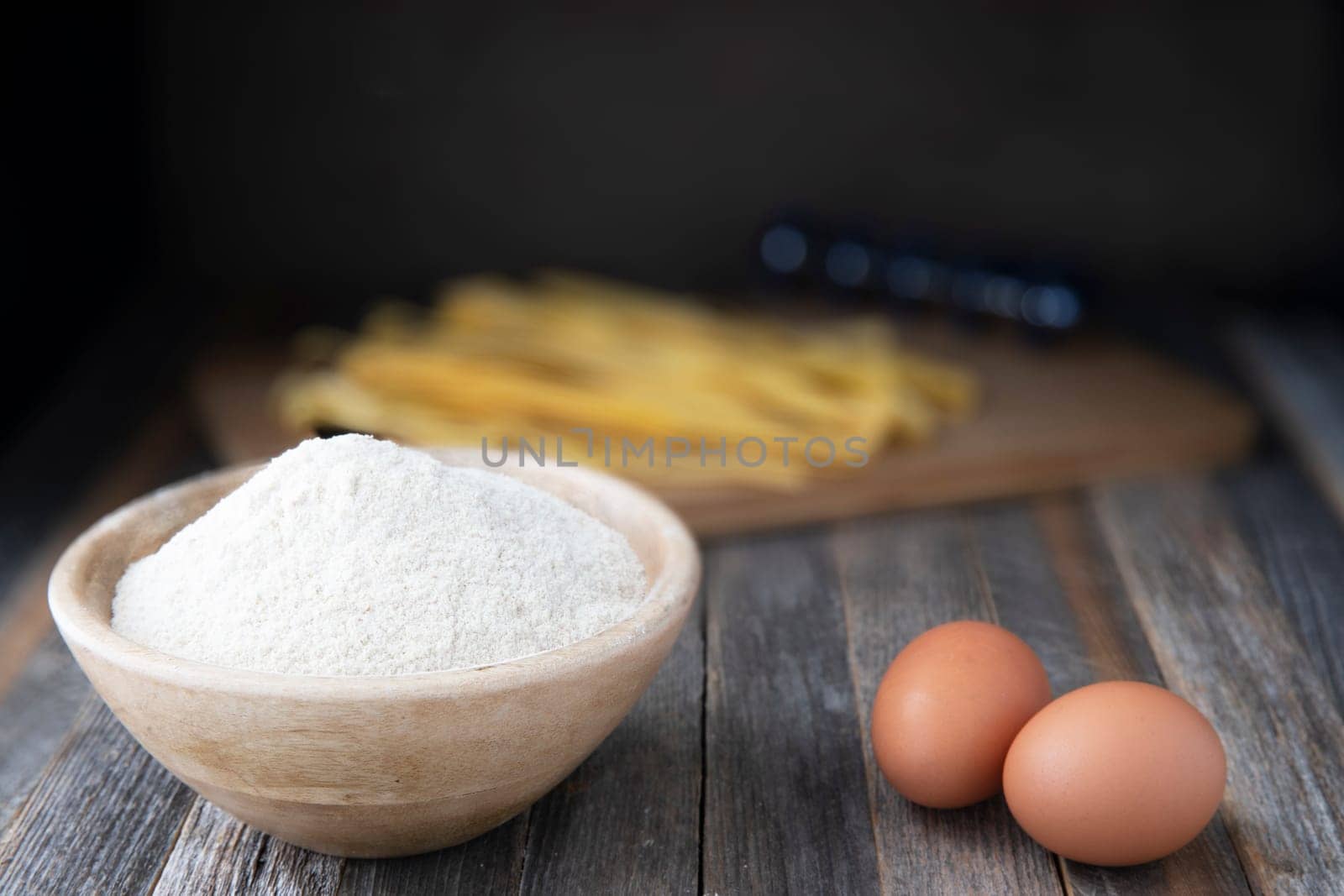 Bowl of whole wheat flour and two eggs on a rustic wooden table with pappardelle pasta in the background.