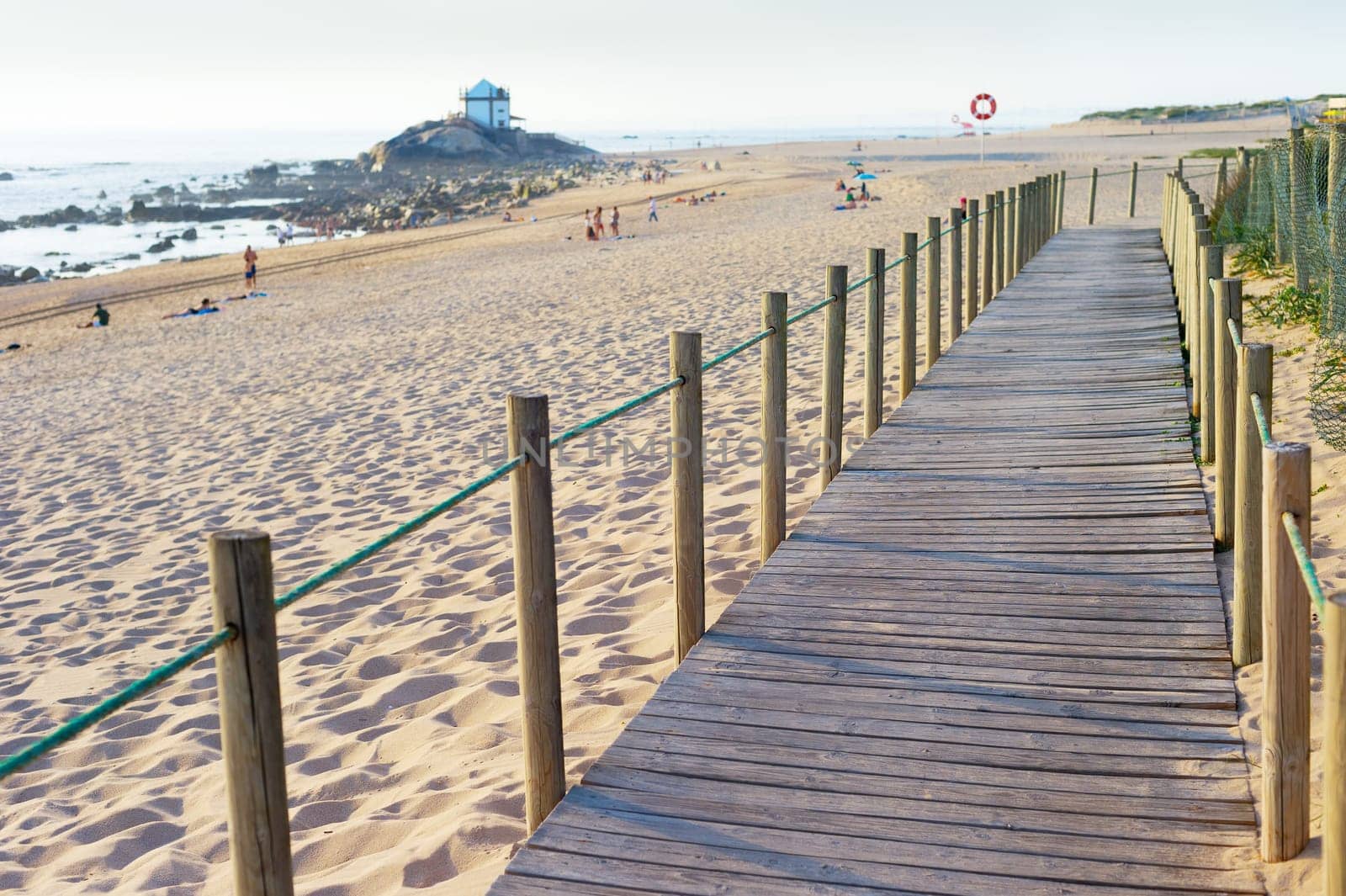 Wooden walkway, people at sandy beach in evening sunlight, Senhor da Pedra church in background by seashore, Porto, Portugal