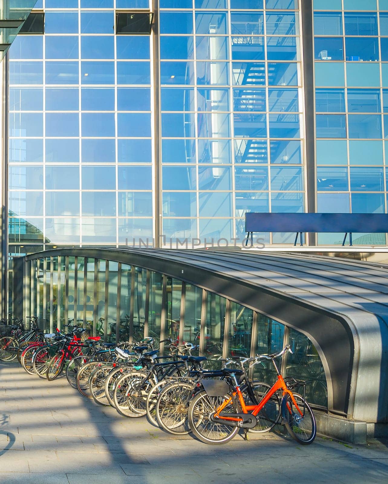 Parking of bicycles by Kastrup airport entrance under canopy, glass building facade in evening sunshine, Copenhagen, Denmark