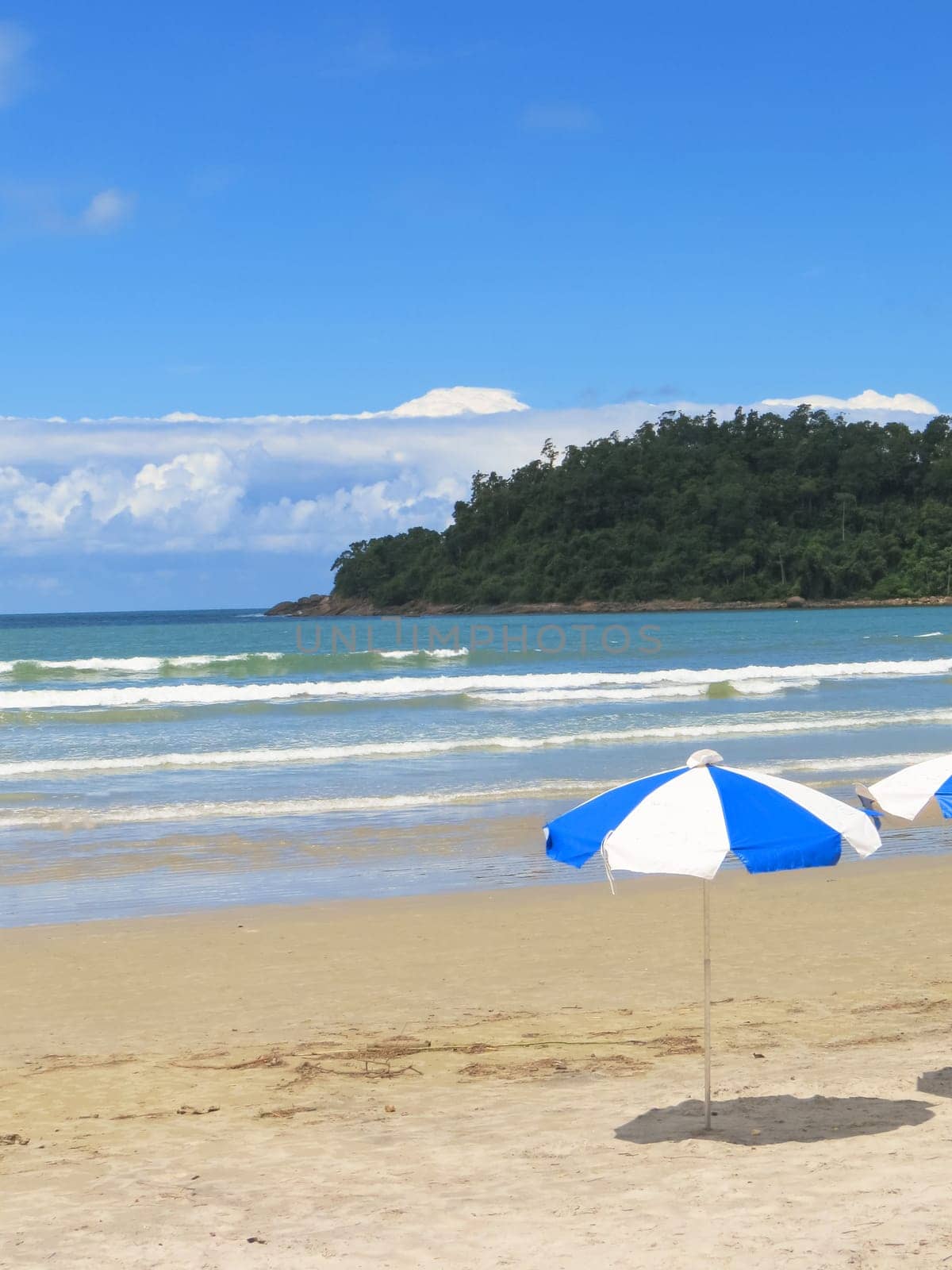 View of sunny beach with umbrella, waves and island in the background.