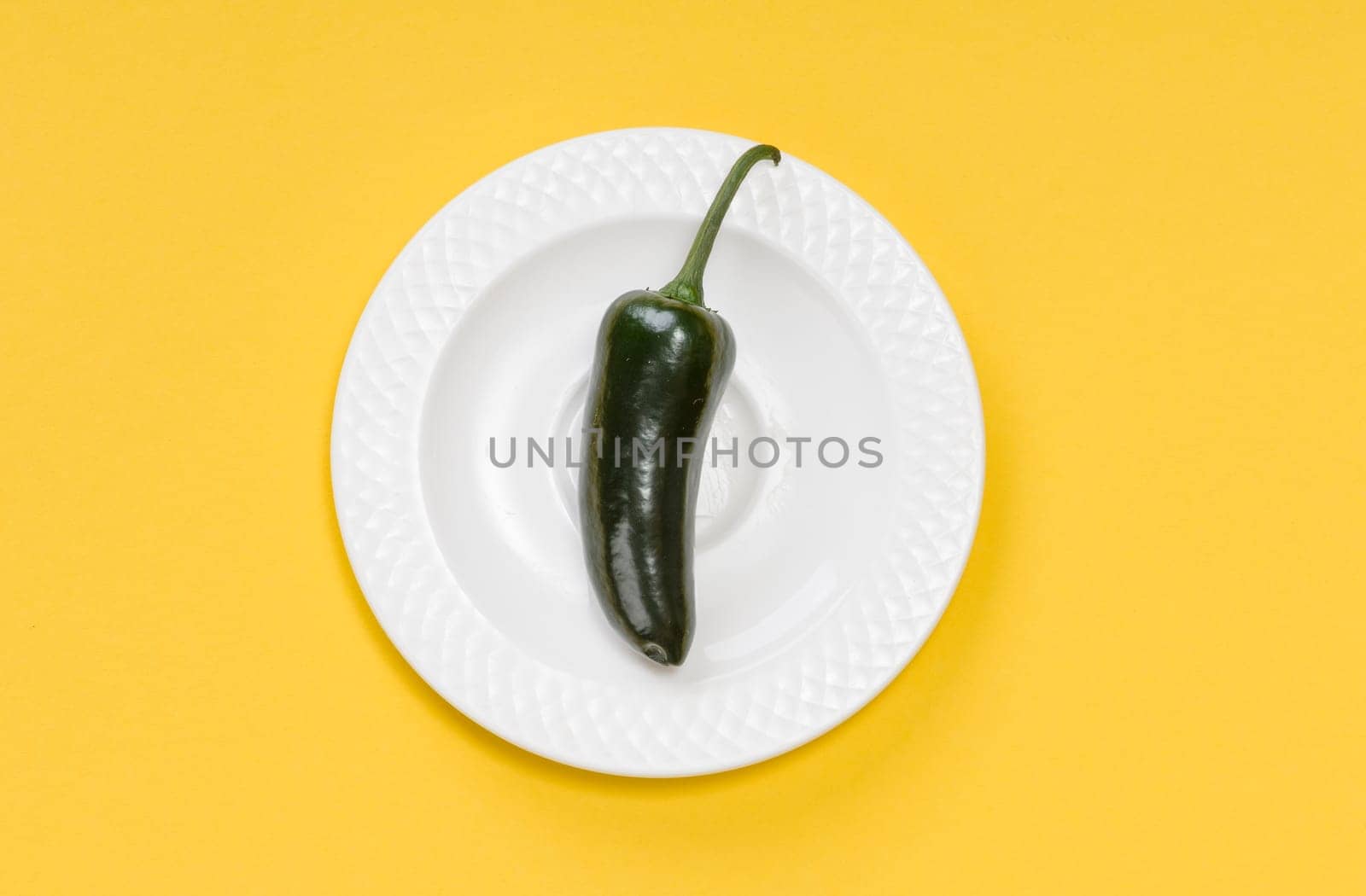 Single green jalapeno pepper on white plate on yellow background