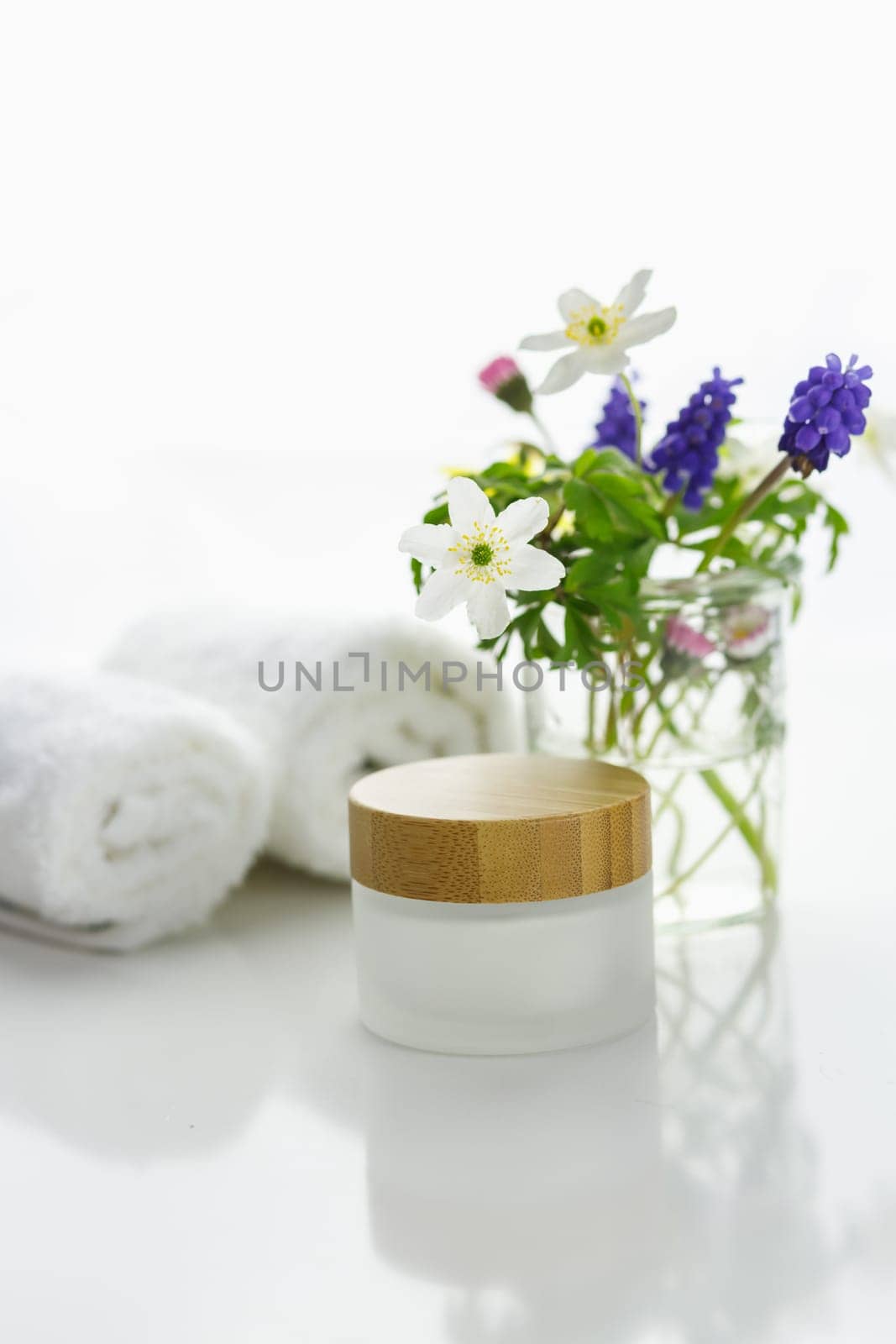 Towels next to a cream jar in front of white background and flowers in a vase.
