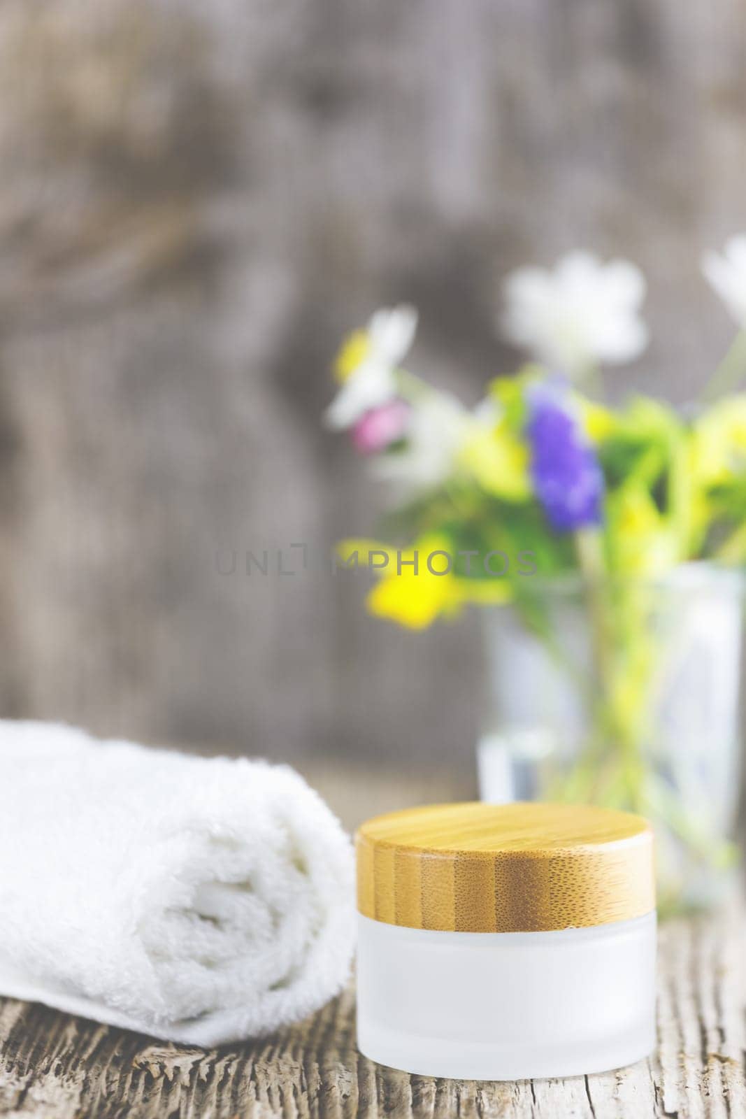 Towels next to a cream jar in front of white background and flowers in a vase.