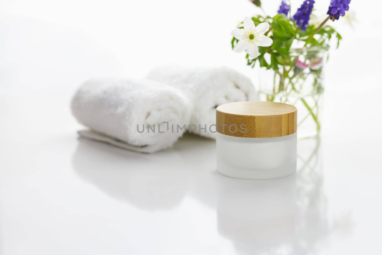 Towels next to a cream jar in front of white background and flowers in a vase.
