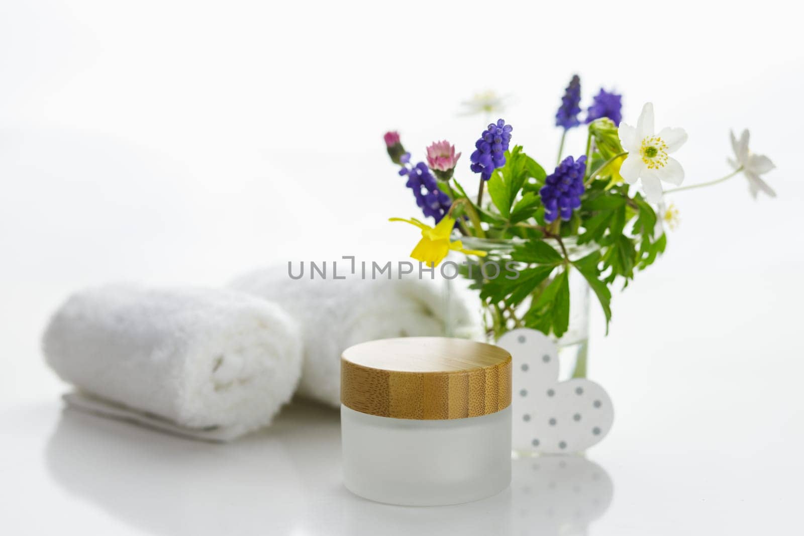 Towels next to a cream jar in front of white background and flowers in a vase.