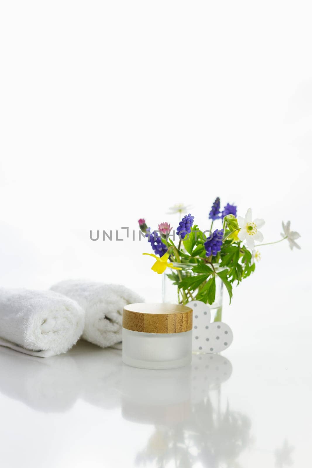 Towels next to a cream jar in front of white background and flowers in a vase.