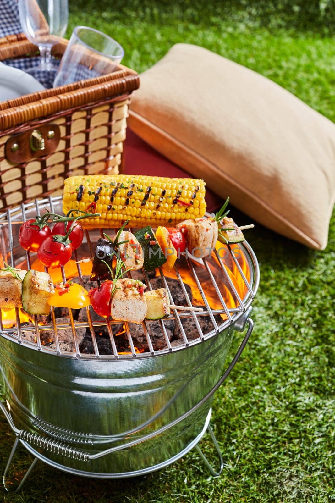 Fresh corn and kebabs with meat and vegetables being grilling on shiny metal bucket grille on green lawn outdoors. Picnic stuff is in background