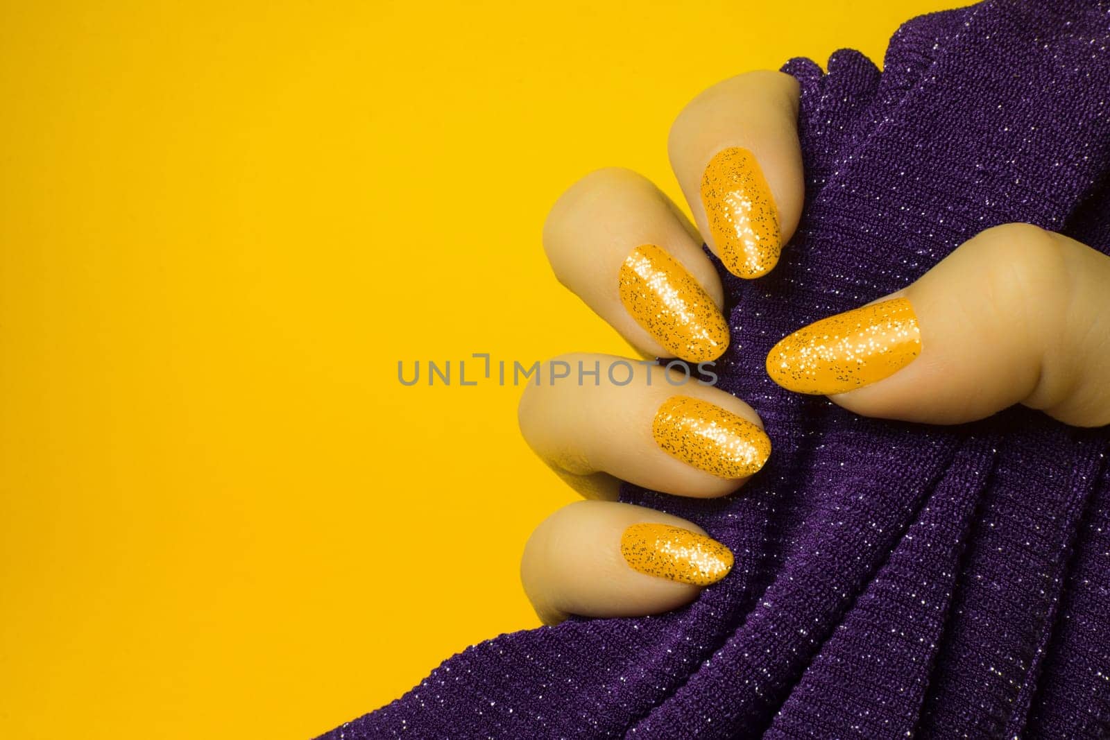 Female hand with glittered yellow nails is holding purple textile on yellow background, manicure and nail care concept.