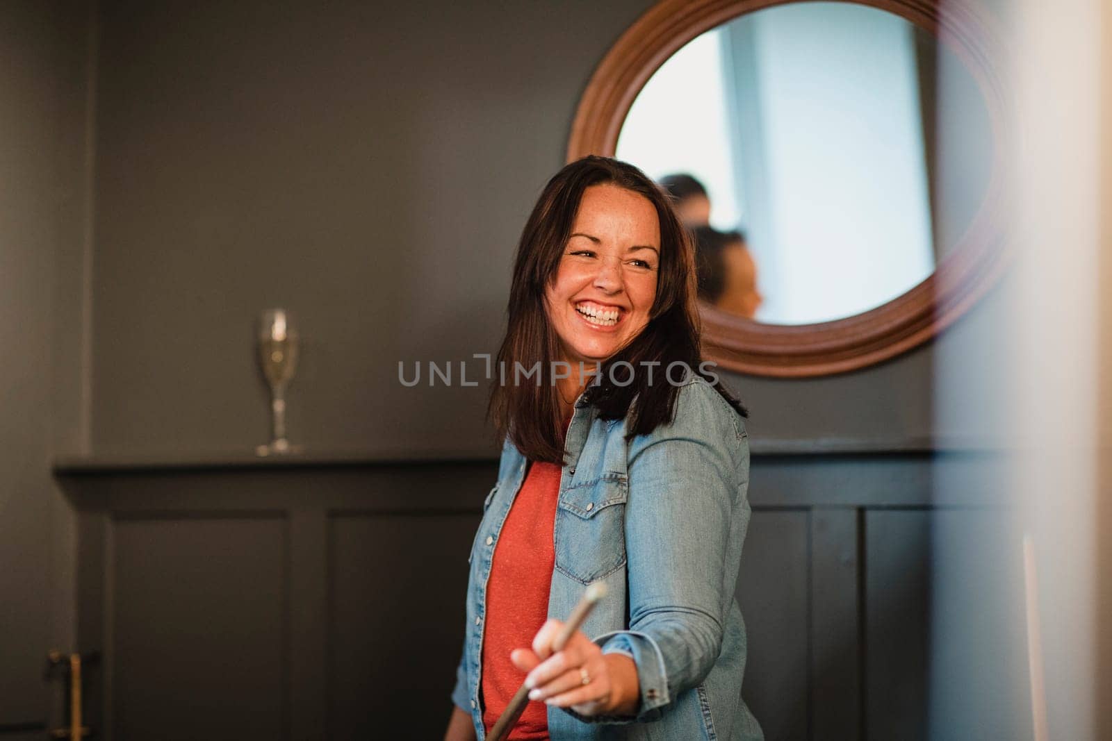 Portrait of a smiling mid adult woman, she is standing indoors holding a pool cue and laughing.