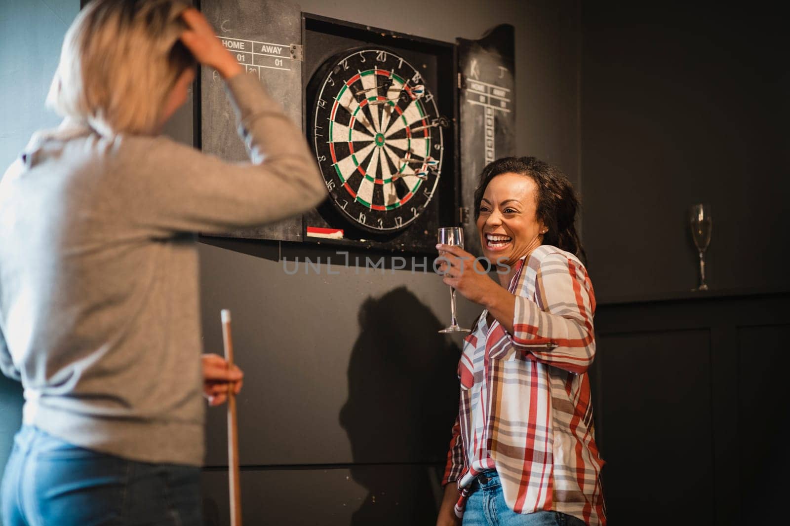 Two female friends relaxing and having fun. They are standing in a games room next to a darts board, enjoying a drink.