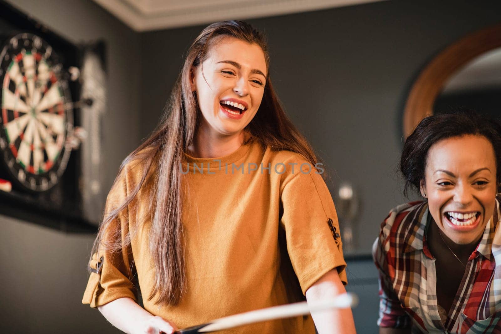 Young female adult having fun and laughing while playing a game of pool with her friend.