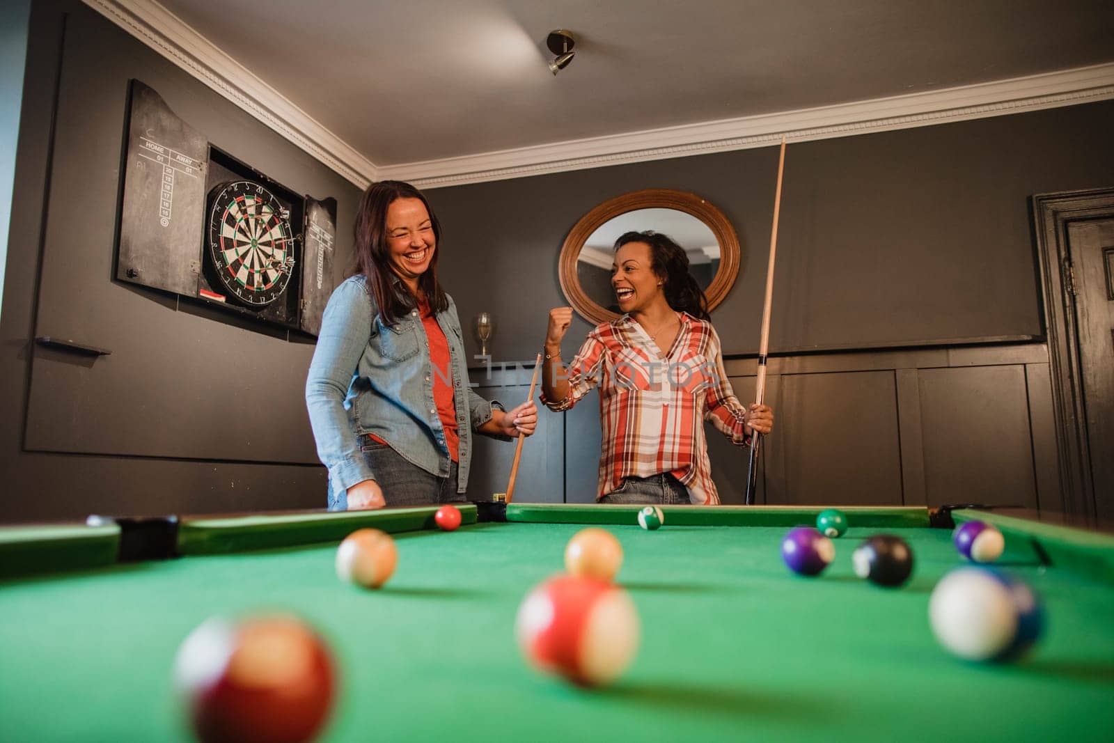 Two mid adult friends playing a game of pool in a games room in a house.