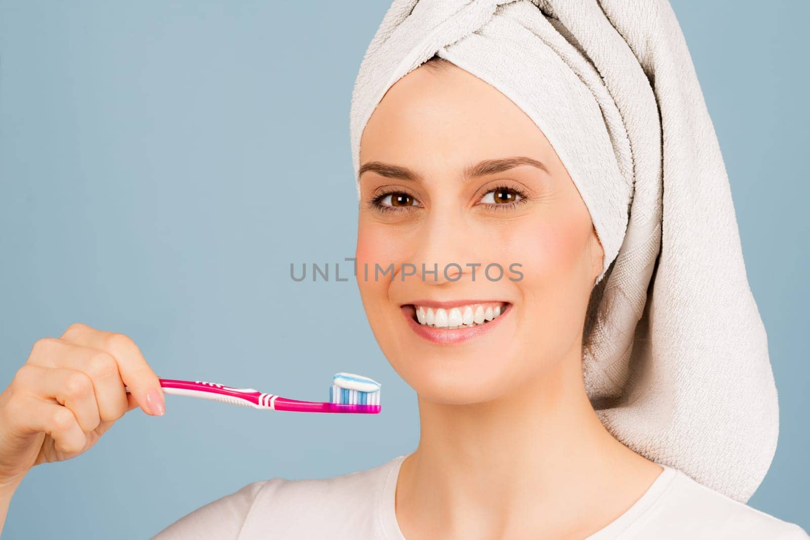 The beautiful smiling woman with white towel on head is ready to brushing teeth on light blue background.