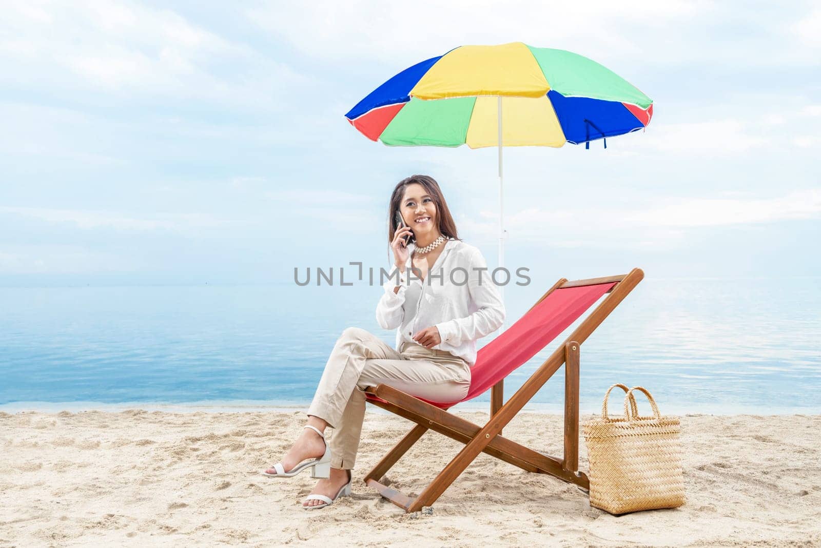 Asian business woman talking on mobile phone while sitting sitting in the beach chair with colorful umbrella on beach. Summer vacation