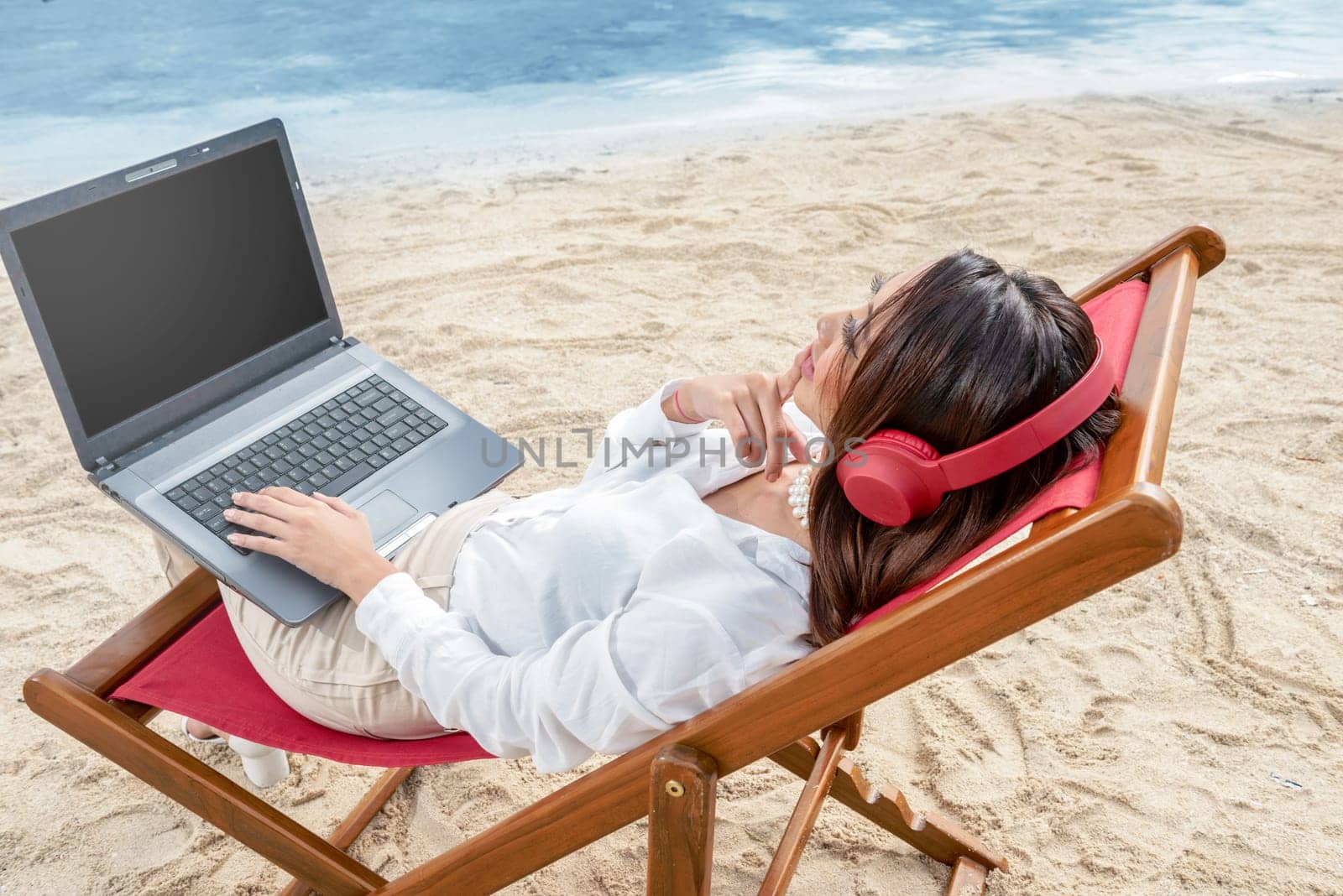 Rear view of asian business woman working with laptop while using headphones sitting in the beach chair and thinking something on beach. Summer vacation
