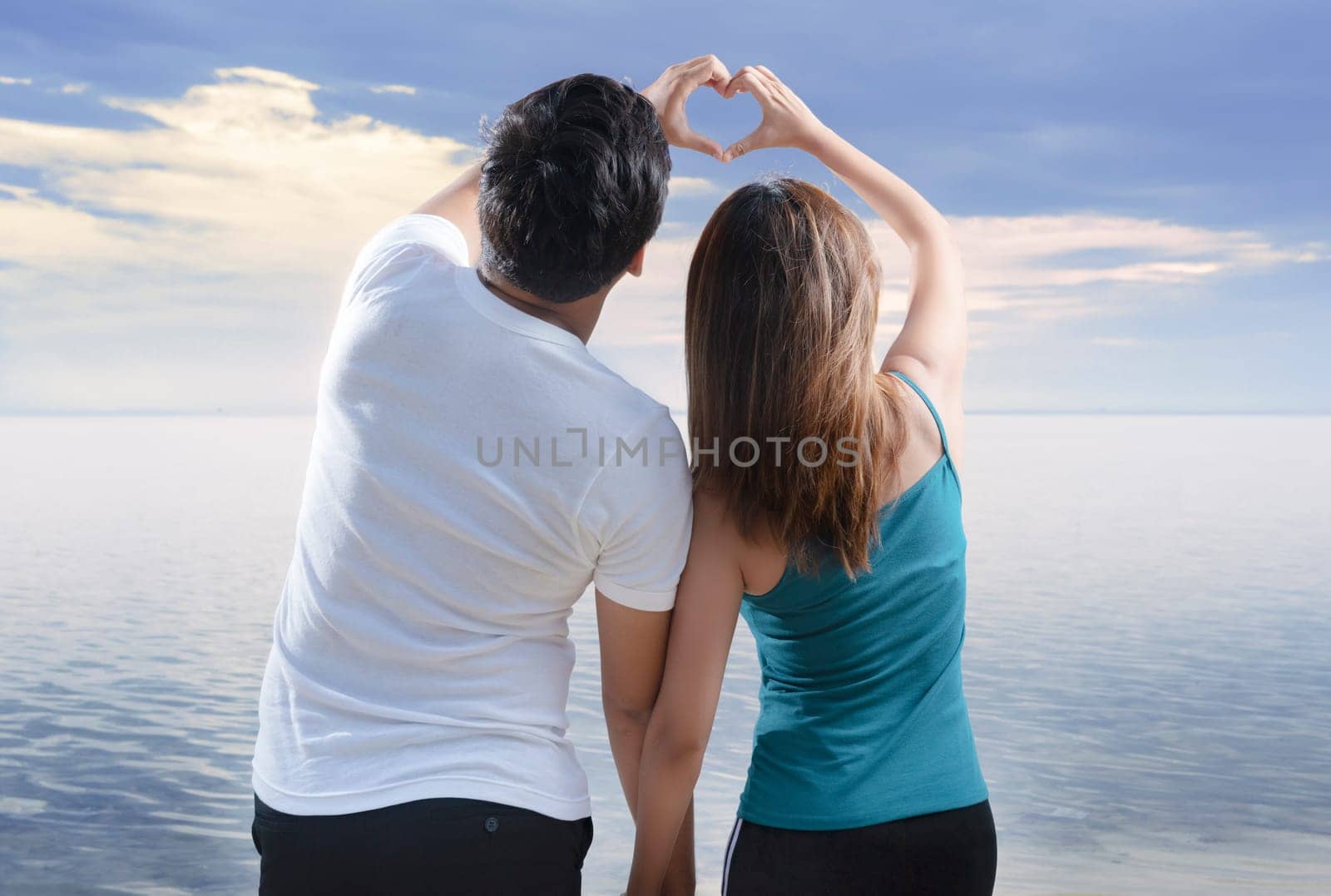 Rear view of asian romantic couple making heart shapes with fingers on the beach. Summer vacation