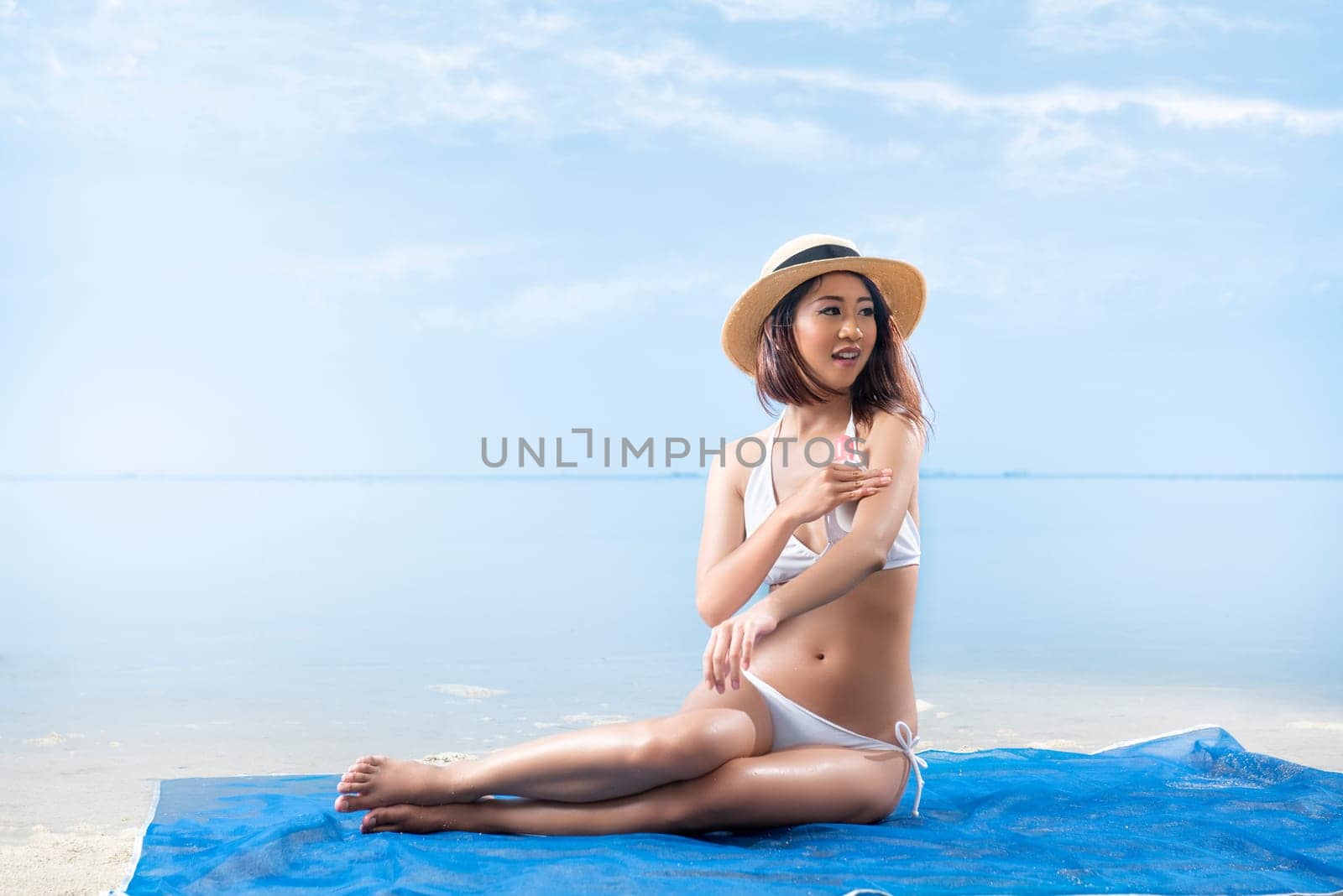 Asian sexy girl in bikini and hat sitting on the carpet and applying oil onto skin in beach. Summer vacation