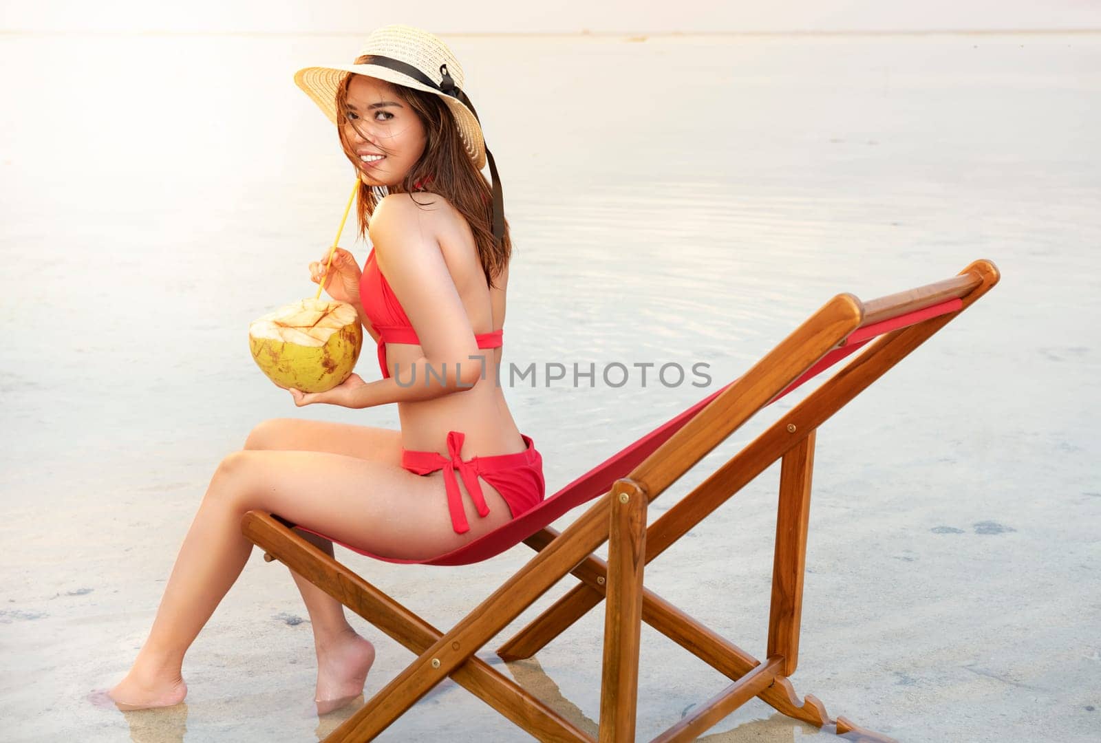 Asian sexy girl in bikini and hat with coconut fruit while sitting on the beach chair at seaside. Summer vacation